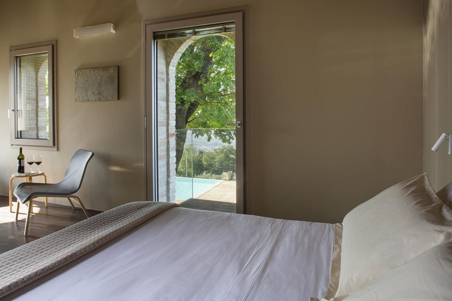Cozy bedroom with a large bed, featuring natural light streaming in through a sliding glass door that opens to a balcony with a view of greenery. A stylish chair and a small table complement the modern decor. | Only on SopranoVillas