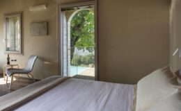 Cozy bedroom with a large bed, featuring natural light streaming in through a sliding glass door that opens to a balcony with a view of greenery. A stylish chair and a small table complement the modern decor.