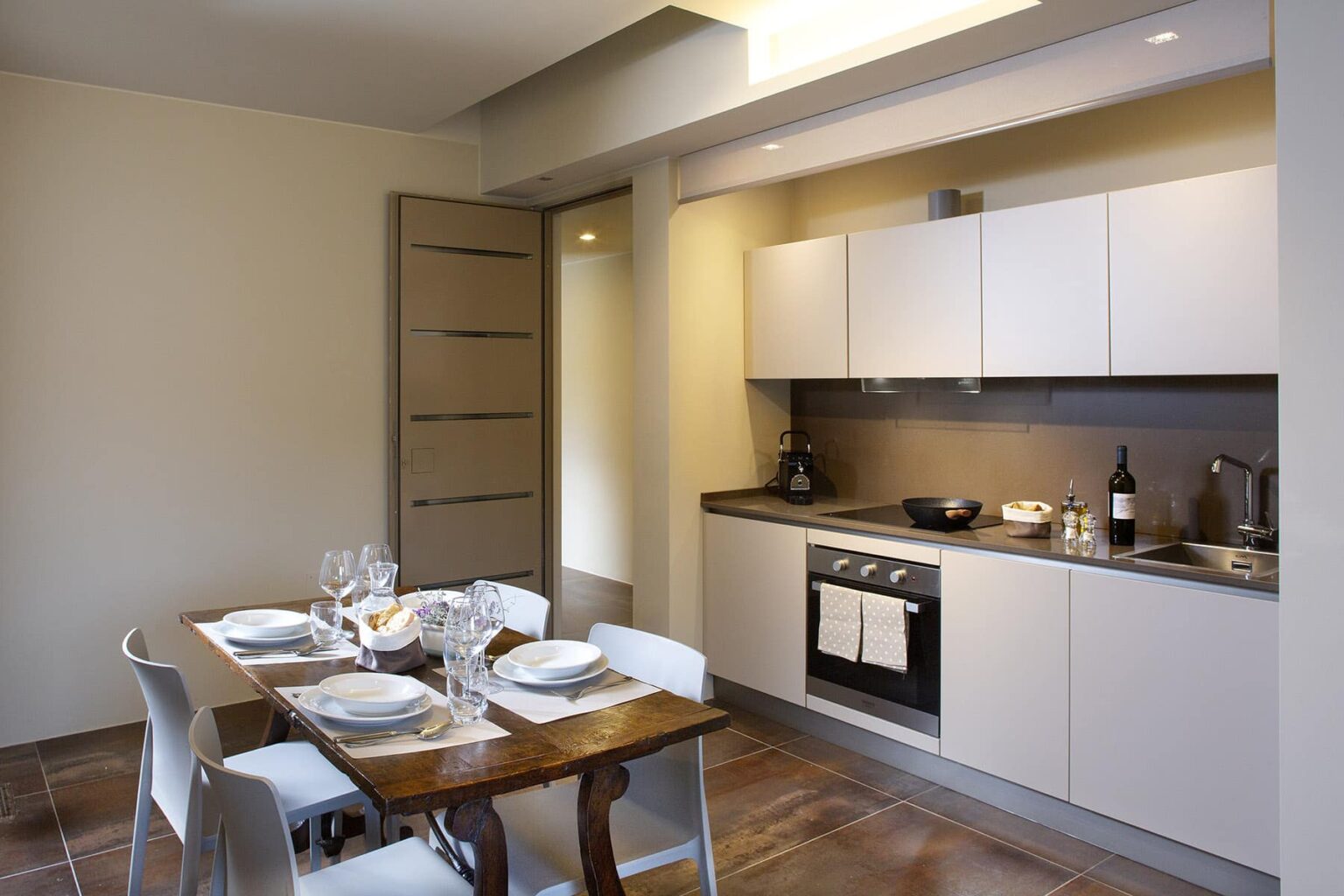 Modern kitchen interior featuring sleek white cabinetry, a dark countertop, and a dining table set for four. The space is well-lit with natural light, showcasing a minimalist design and contemporary appliances. | Only on SopranoVillas
