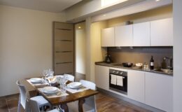 Modern kitchen interior featuring sleek white cabinetry, a dark countertop, and a dining table set for four. The space is well-lit with natural light, showcasing a minimalist design and contemporary appliances.