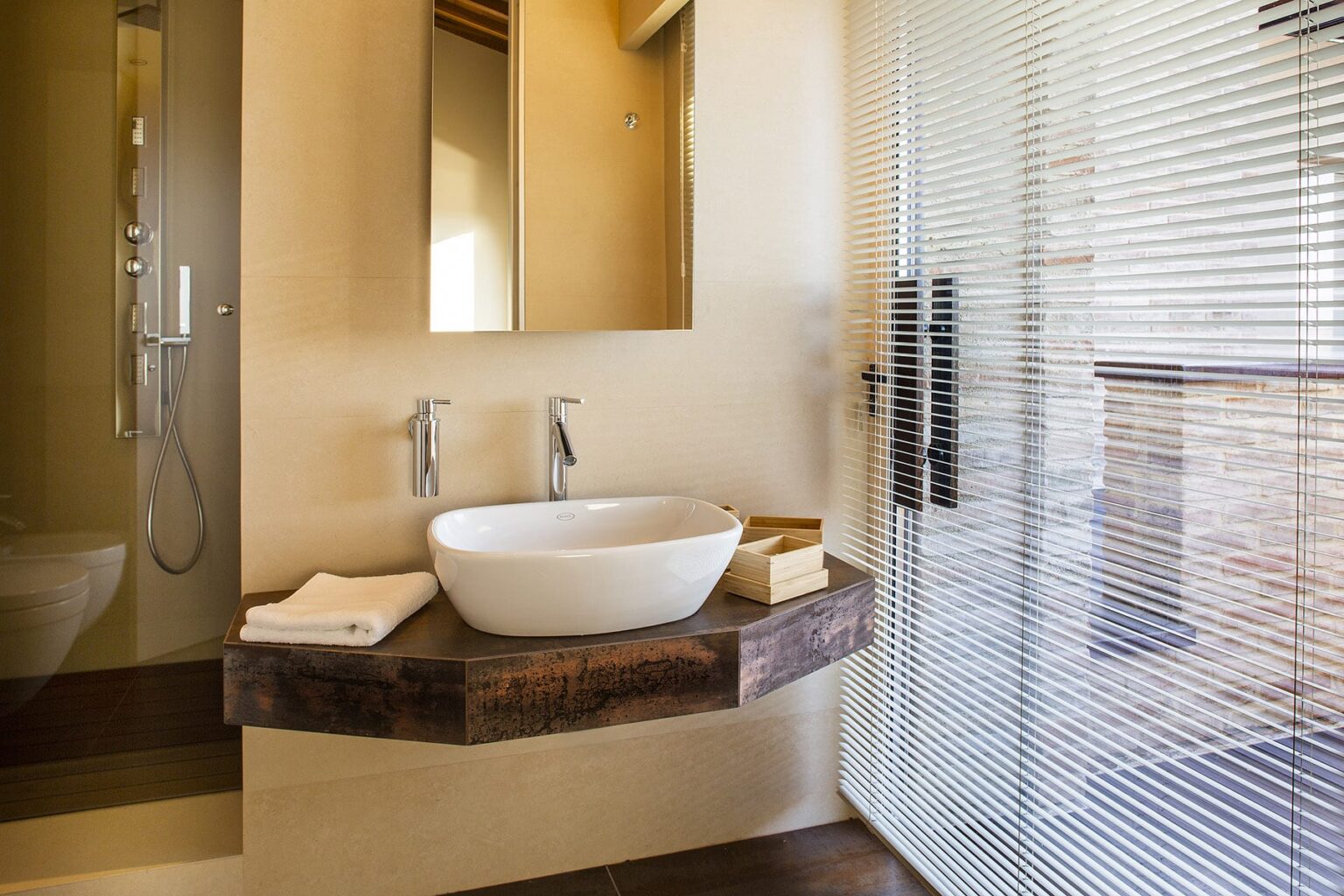 Modern bathroom featuring a stylish vessel sink on a wooden countertop, sleek faucets, a large mirror, and natural light from a glass door with blinds. | Only on SopranoVillas