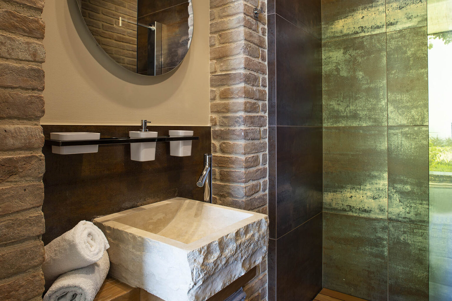 Modern bathroom interior featuring a stone sink, round mirror, and a glass shower. The walls are adorned with textured stone, creating a warm and inviting atmosphere. White towels are neatly arranged, enhancing the overall aesthetic. | Only on SopranoVillas