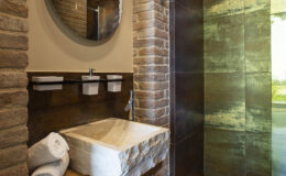 Modern bathroom interior featuring a stone sink, round mirror, and a glass shower. The walls are adorned with textured stone, creating a warm and inviting atmosphere. White towels are neatly arranged, enhancing the overall aesthetic.