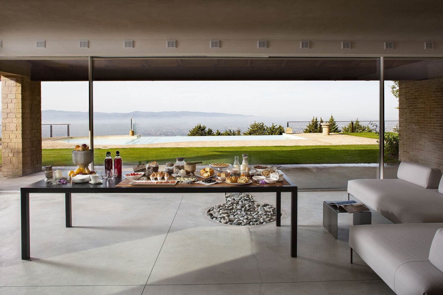 Modern outdoor dining area featuring a stylish table set with an array of gourmet food and beverages, overlooking a scenic landscape with a swimming pool and lush greenery. Ideal for entertaining guests in a luxurious setting. | Only on SopranoVillas