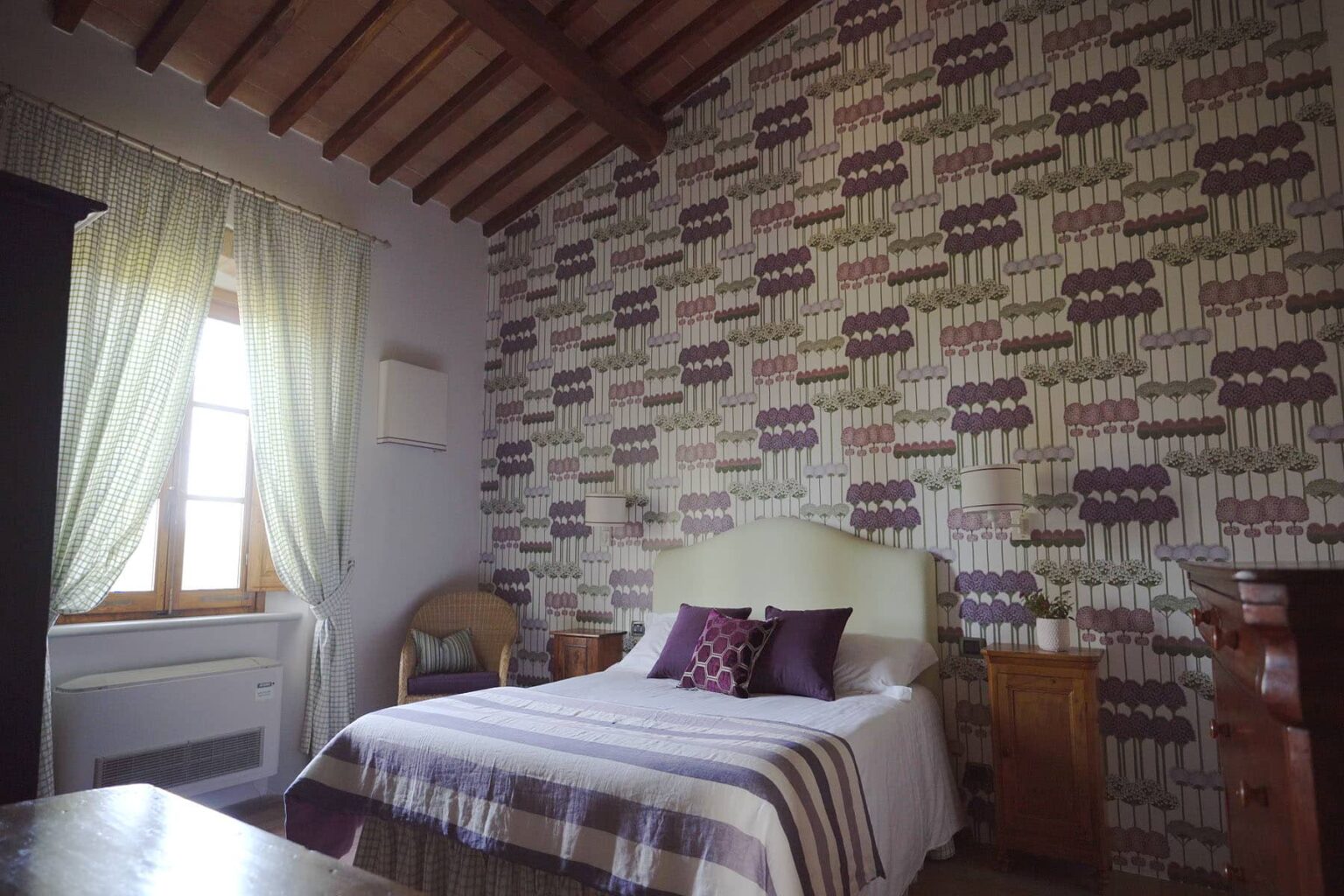 Cozy bedroom featuring a stylish patterned wallpaper, a comfortable bed with decorative pillows, wooden ceiling beams, and natural light streaming through sheer curtains. | Only on SopranoVillas