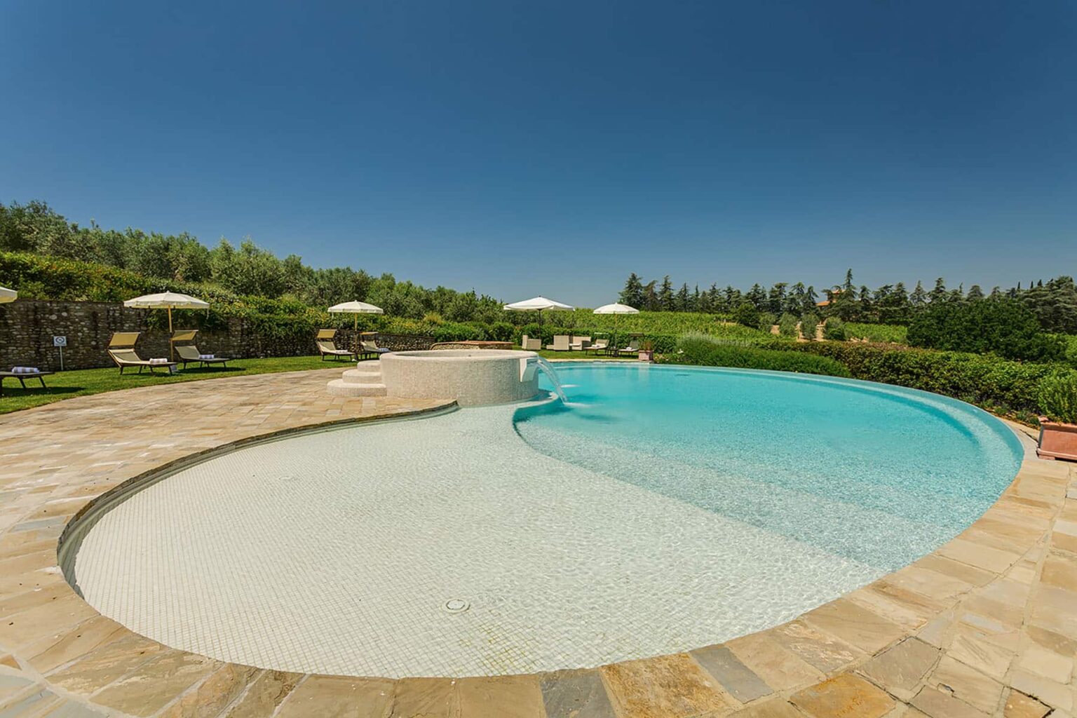 Modern outdoor swimming pool surrounded by lush greenery and sun loungers under umbrellas, set against a clear blue sky. Ideal for luxury vacation settings or resort imagery. | Only on SopranoVillas