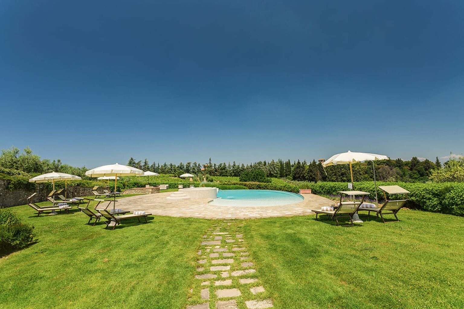 Scenic outdoor pool area surrounded by lush greenery, featuring lounge chairs and umbrellas under a clear blue sky. Perfect for relaxation and leisure activities. | Only on SopranoVillas