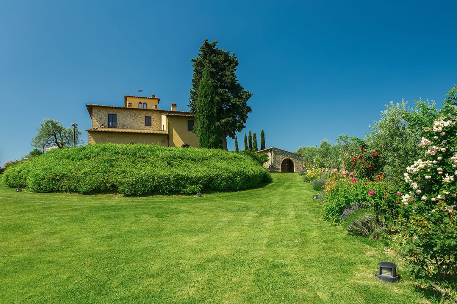 Scenic view of a charming hillside villa surrounded by lush green gardens and trees under a clear blue sky. The landscape features well-maintained grass and flowering shrubs, creating a picturesque outdoor setting. | Only on SopranoVillas