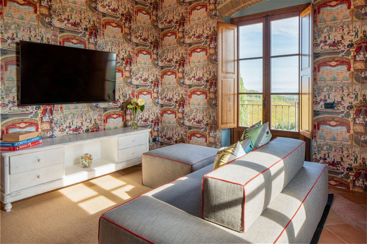 Cozy living room featuring a patterned wallpaper, a modern white TV stand, a flat-screen television, and a stylish gray sofa with red accents, illuminated by natural light from large windows. | Only on SopranoVillas