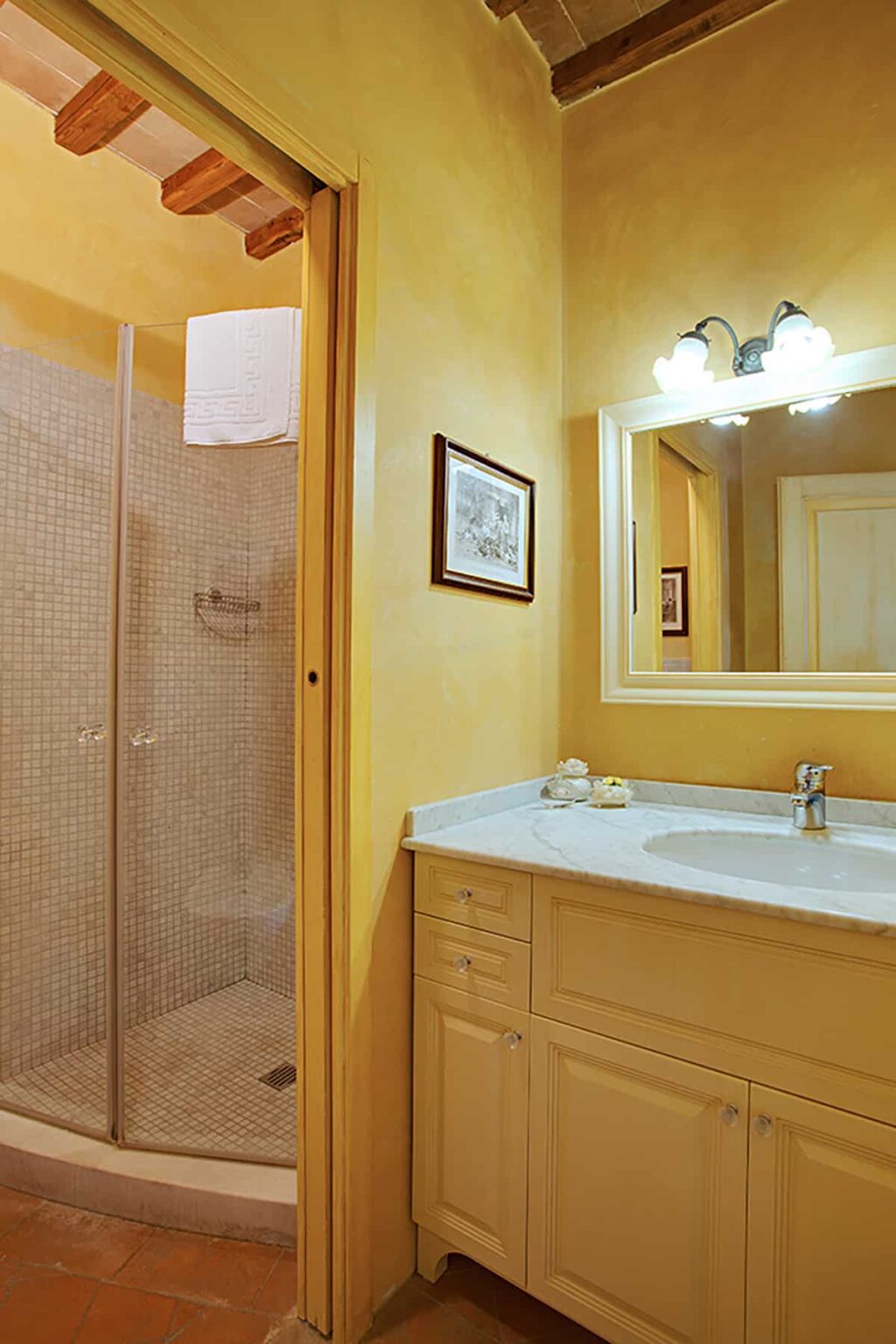 Bright and inviting bathroom featuring a marble countertop, a large mirror, and a walk-in shower with mosaic tiles. The warm yellow walls enhance the cozy atmosphere, complemented by decorative artwork. | Only on SopranoVillas