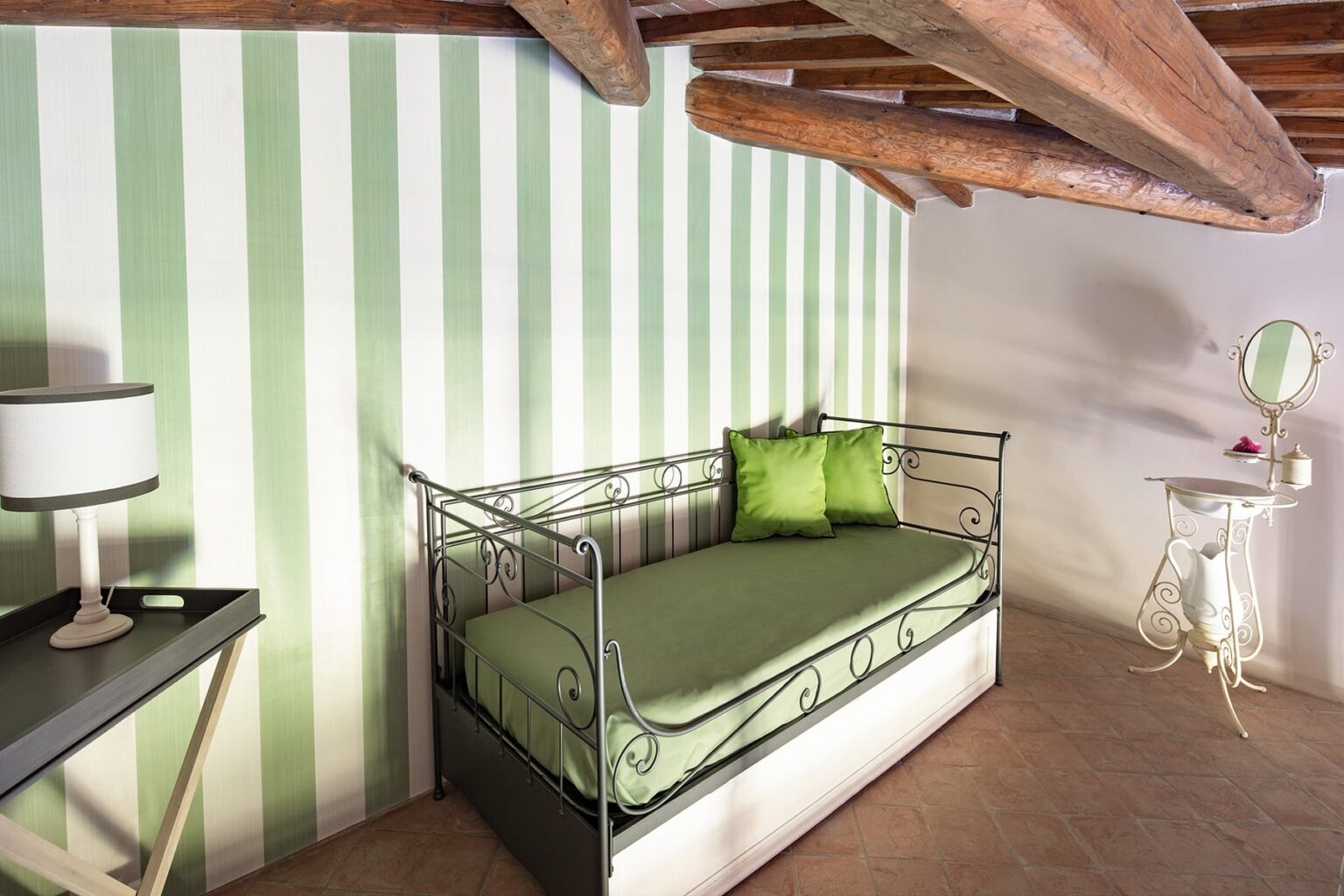 Cozy interior of a room featuring a green and white striped wall, a metal daybed with green cushions, a small side table, and a vintage mirror, showcasing a charming and inviting atmosphere. | Only on SopranoVillas