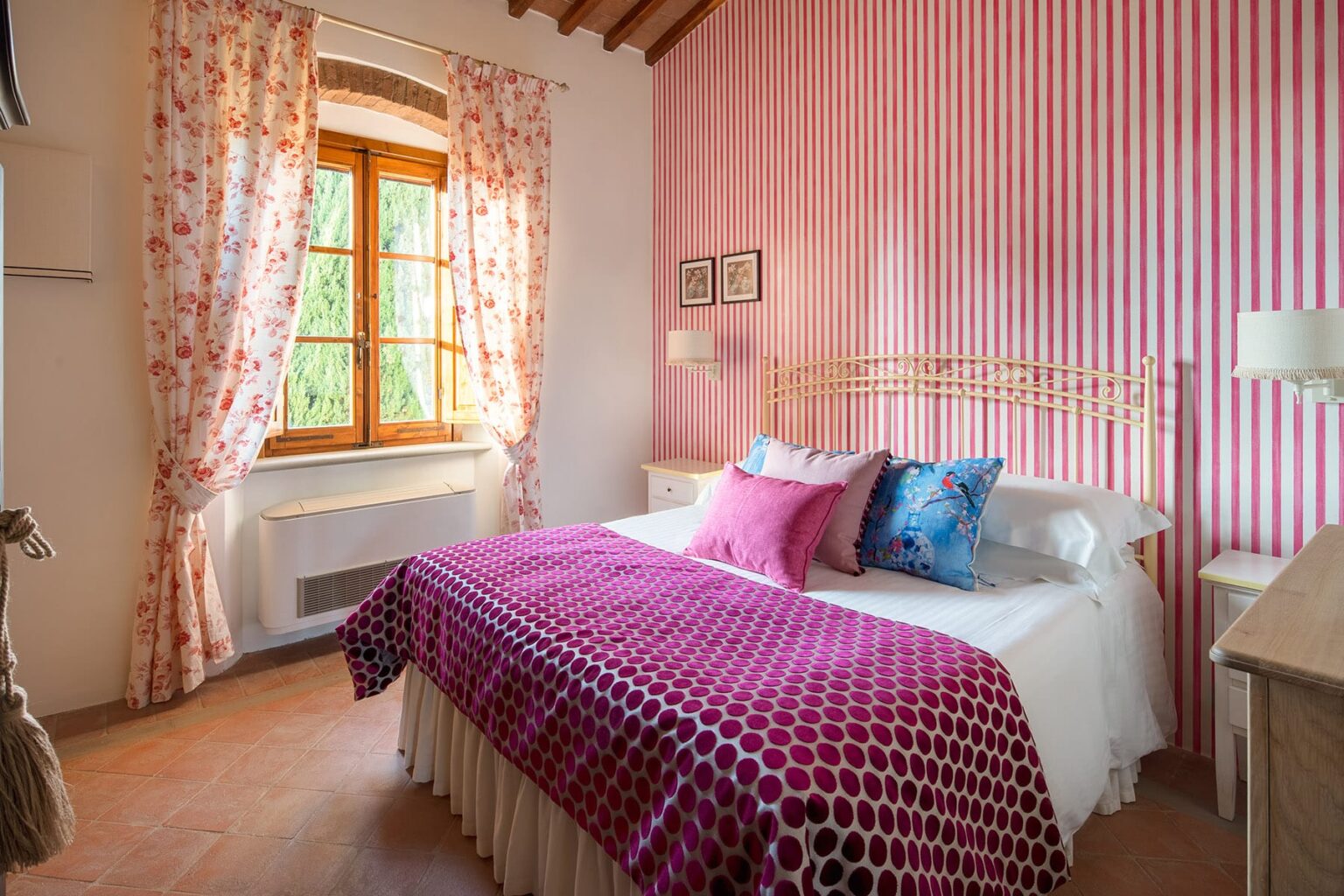 Cozy bedroom featuring a queen-sized bed with colorful pillows, floral curtains, and pink striped walls, illuminated by natural light from a window. Ideal for a relaxing getaway or home decor inspiration. | Only on SopranoVillas
