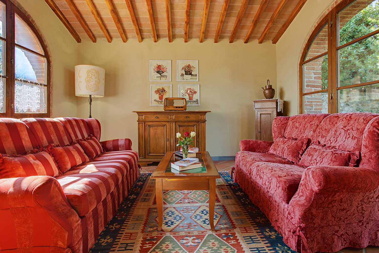 Cozy living room featuring two red sofas, a wooden coffee table, and decorative artwork on the walls. Natural light streams in through large windows, highlighting the rustic wooden ceiling and vibrant area rug. Ideal for relaxation and gatherings. | Only on SopranoVillas
