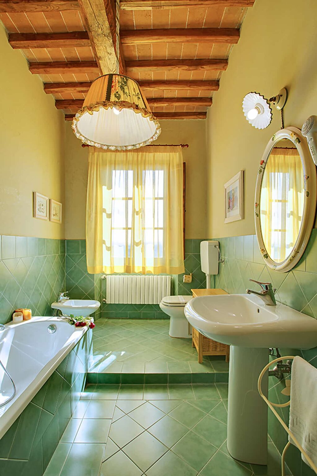 Bright and airy bathroom featuring a large window with yellow curtains, a round mirror, a sink, a toilet, and a bidet, complemented by green tiled walls and wooden accents. | Only on SopranoVillas