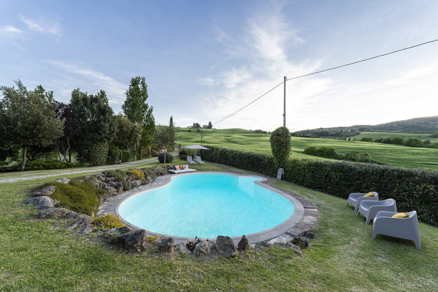Scenic outdoor swimming pool surrounded by lush greenery and rolling hills, featuring comfortable lounge chairs and a clear blue sky. Ideal for relaxation and leisure in a tranquil setting. | Only on SopranoVillas