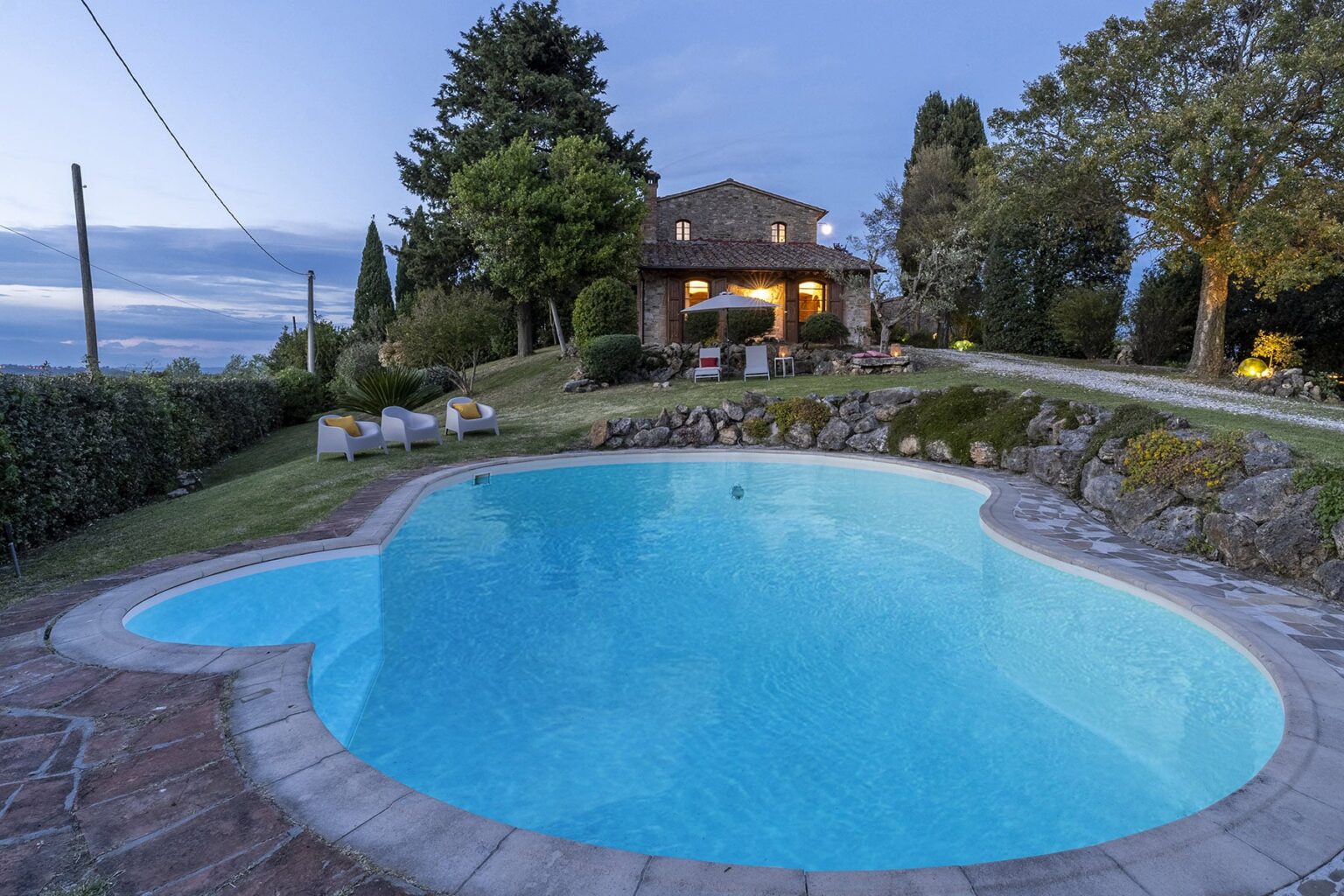Scenic view of a heart-shaped swimming pool surrounded by lush greenery, with a cozy stone house illuminated at dusk in the background. Perfect for a relaxing getaway. | Only on SopranoVillas