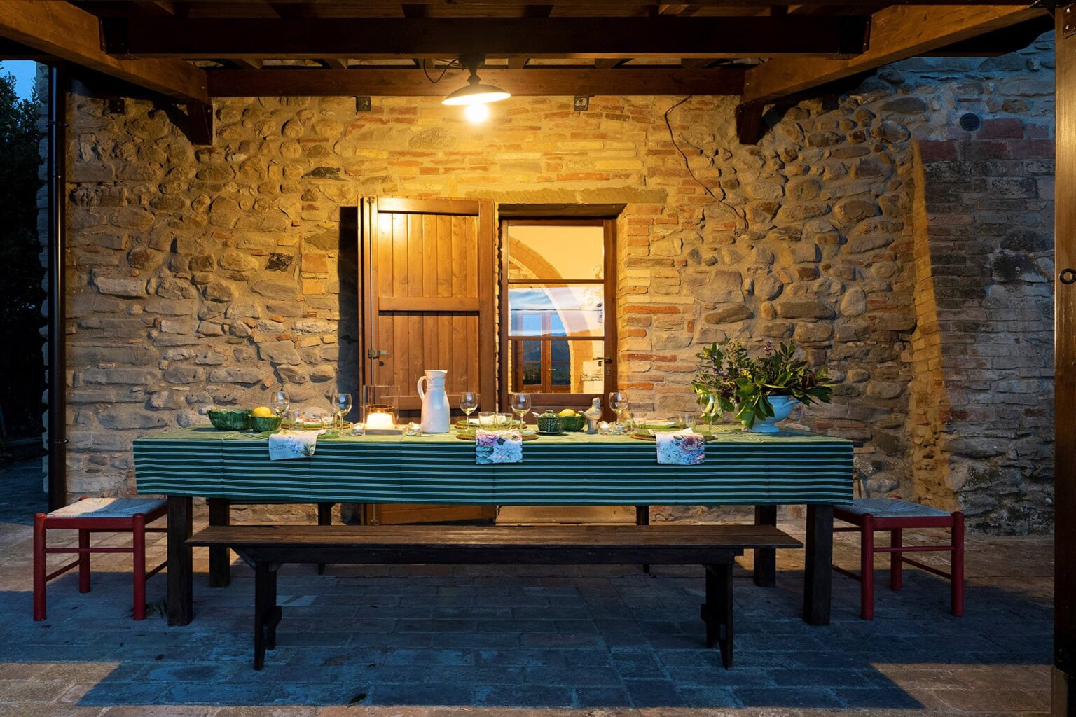 Outdoor dining area with a rustic stone wall, illuminated by warm light, featuring a long wooden table set with tableware and surrounded by red chairs, creating an inviting atmosphere for evening meals. | Only on SopranoVillas