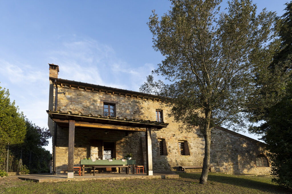 Historic stone farmhouse surrounded by greenery, featuring a spacious porch and large windows under a clear blue sky. Ideal for rural retreats and nature escapes. | Only on SopranoVillas