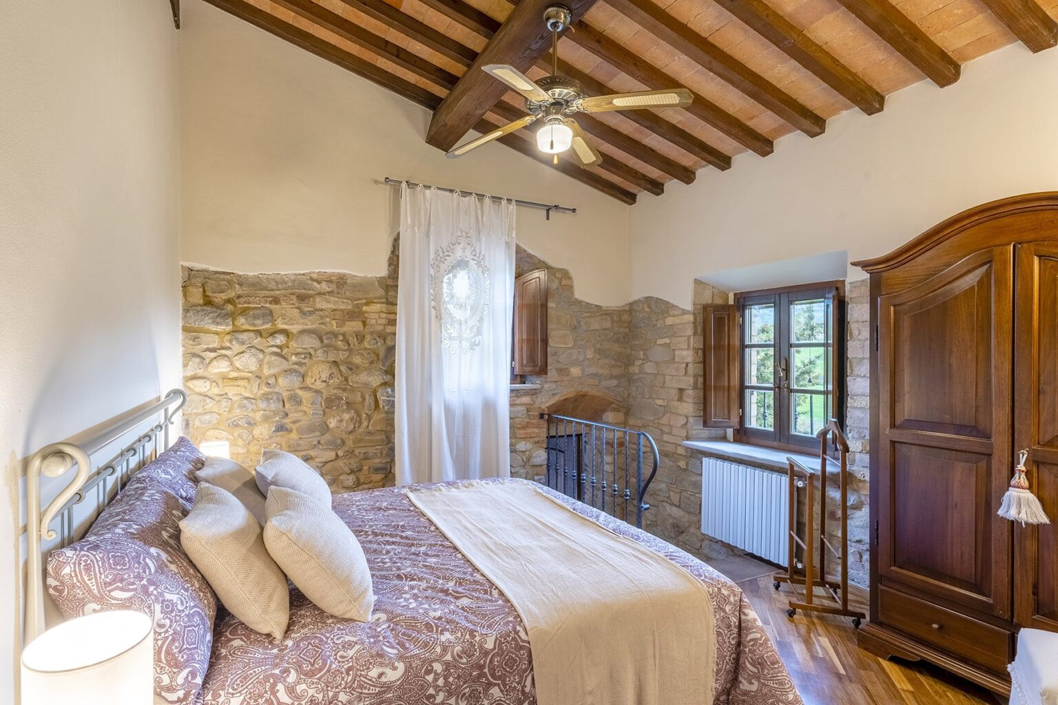 Cozy bedroom featuring a stone wall, wooden ceiling beams, a comfortable bed with decorative pillows, and a window that lets in natural light, creating a warm and inviting atmosphere. | Only on SopranoVillas
