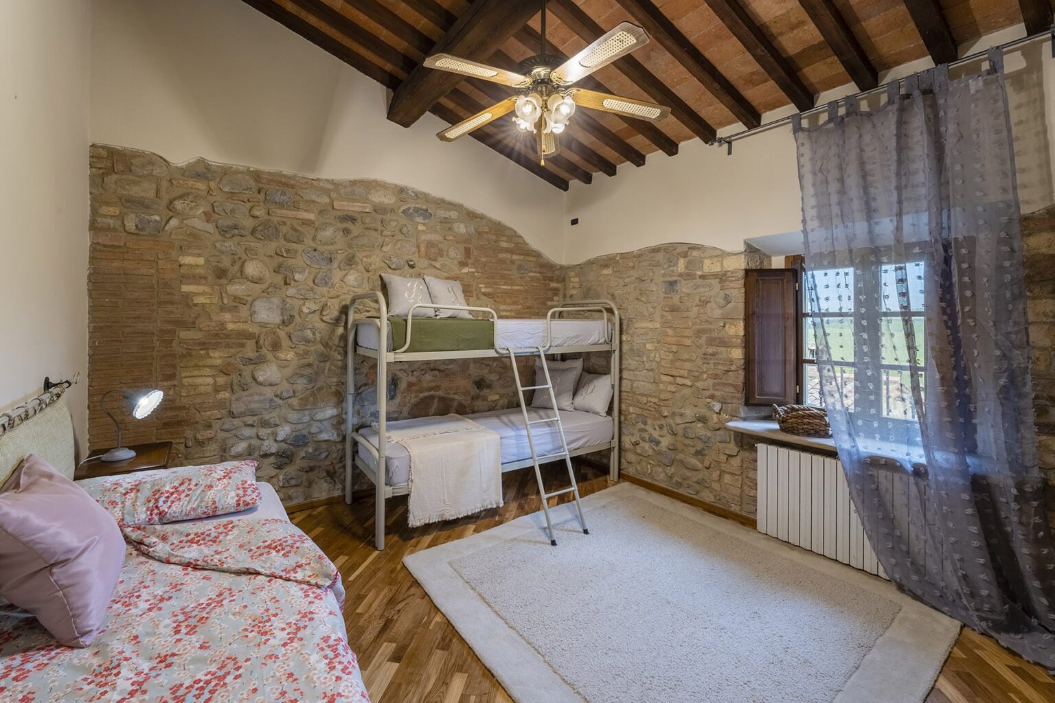 Cozy bedroom featuring a bunk bed, a single bed with floral bedding, exposed stone walls, wooden ceiling beams, and a large window with sheer curtains. Ideal for family stays or group accommodations. | Only on SopranoVillas
