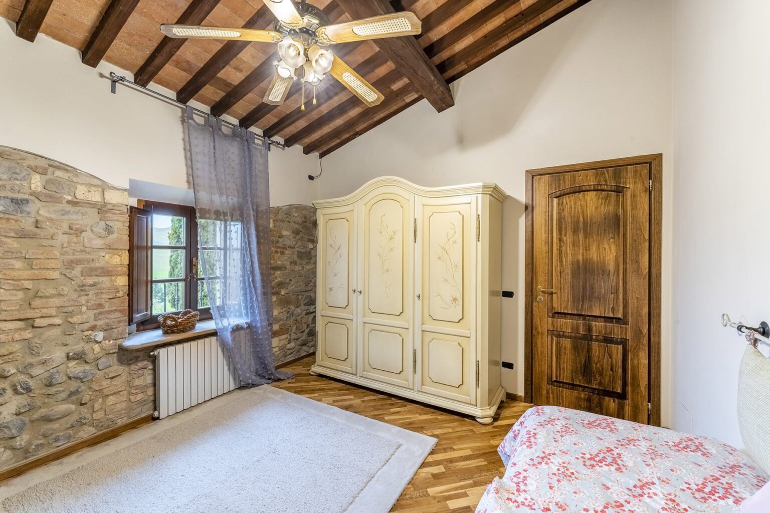 Cozy bedroom interior featuring a wooden ceiling with exposed beams, a vintage white wardrobe, a large window with sheer curtains, and a decorative rug on hardwood flooring. Natural light illuminates the space, creating a warm and inviting atmosphere. | Only on SopranoVillas