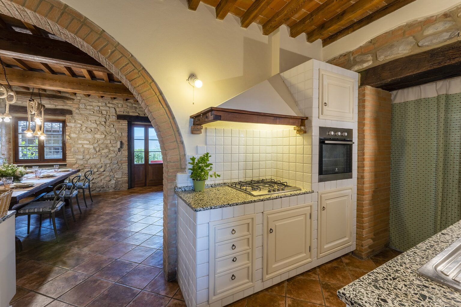 Modern kitchen with granite countertops, white cabinetry, and a built-in oven, featuring an archway leading to a dining area with rustic stone walls and wooden beams. | Only on SopranoVillas