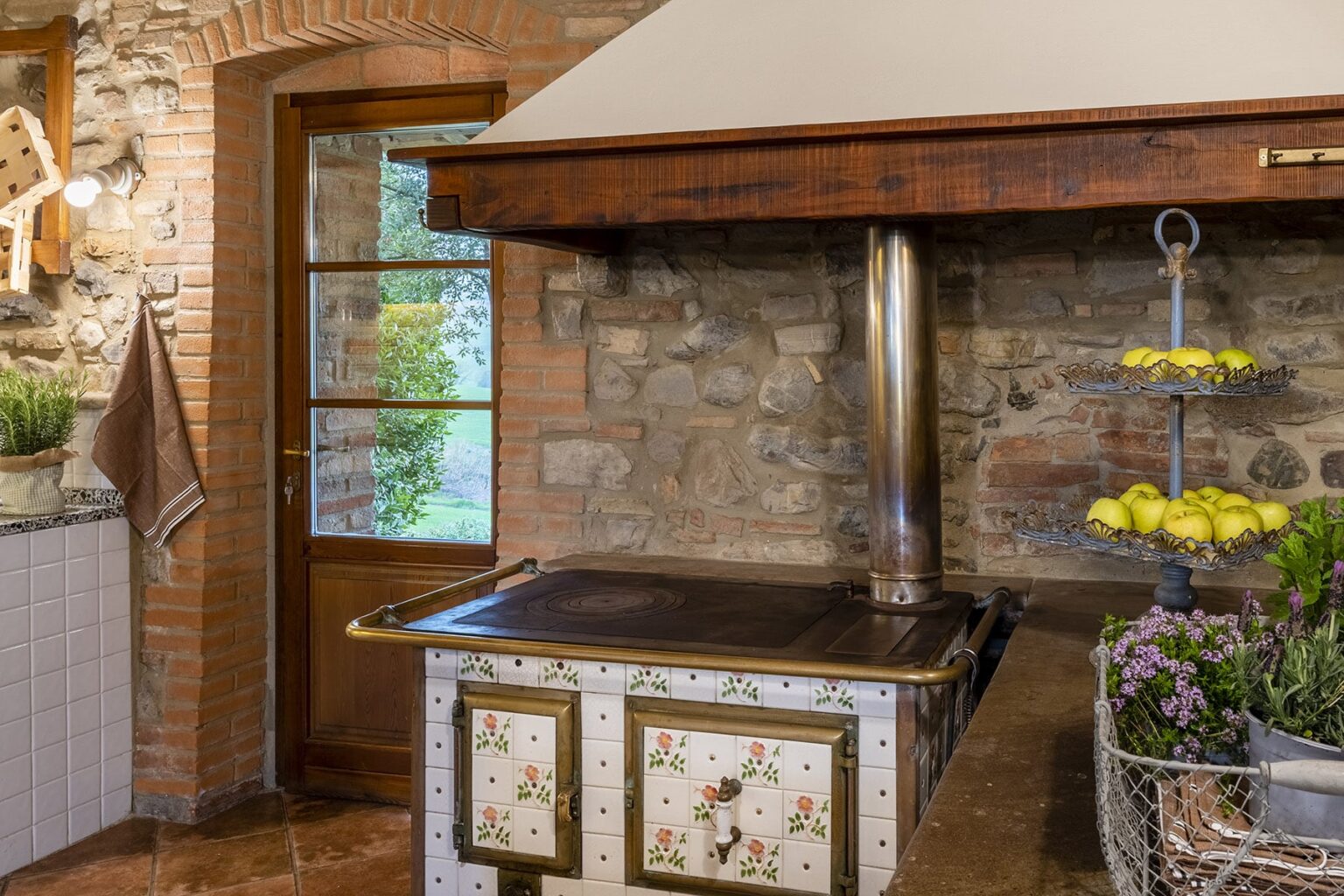 Rustic kitchen interior featuring a decorative stone wall and a vintage wood-burning stove with a metal chimney. The stove is adorned with floral designs, complemented by a nearby fruit display and fresh herbs, creating a warm and inviting atmosphere. | Only on SopranoVillas