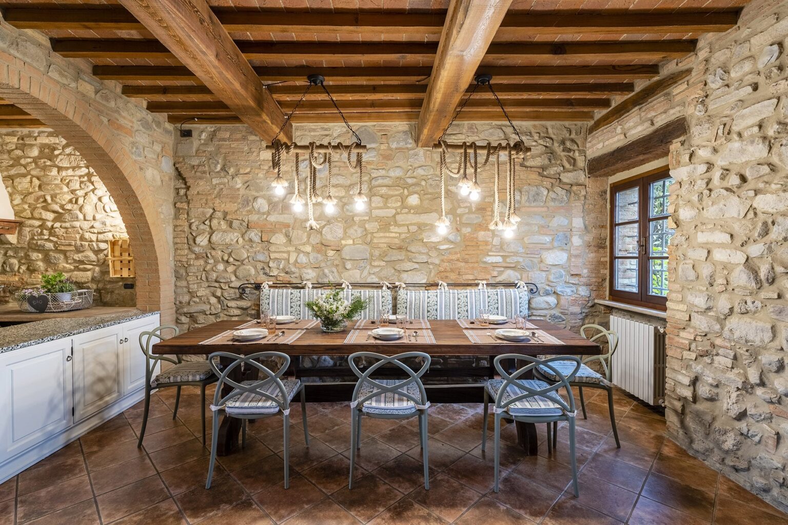 Rustic dining room featuring a large wooden table surrounded by stylish chairs, set against a backdrop of exposed stone walls and wooden beams, with pendant lighting adding warmth to the space. | Only on SopranoVillas
