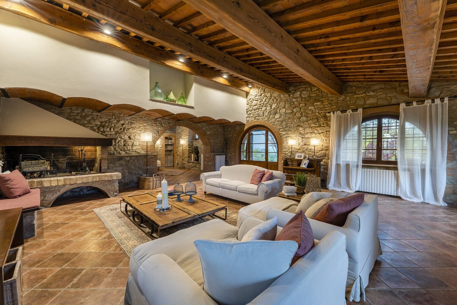 Cozy living room with stone walls, wooden beams, and large windows, featuring comfortable sofas, a coffee table, and a fireplace, creating a warm and inviting atmosphere. | Only on SopranoVillas