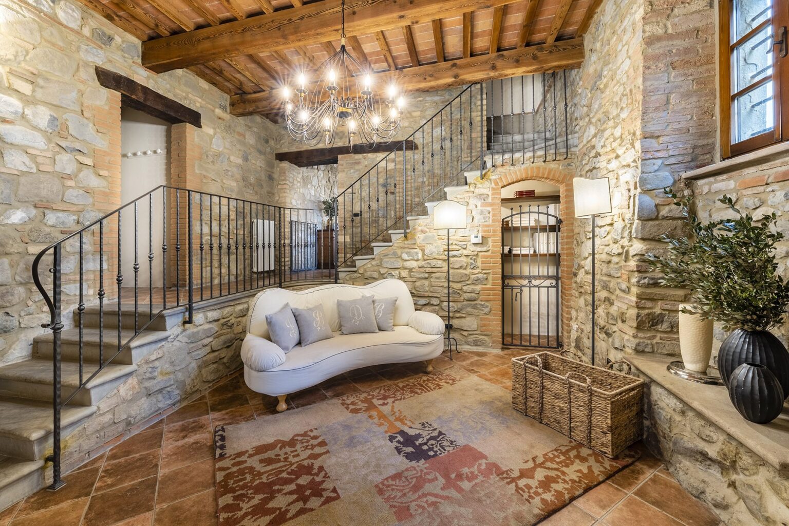 Cozy entrance hall featuring stone walls, wooden ceiling beams, and a stylish staircase. A white sofa and decorative plants enhance the inviting atmosphere, complemented by warm lighting from a chandelier. | Only on SopranoVillas