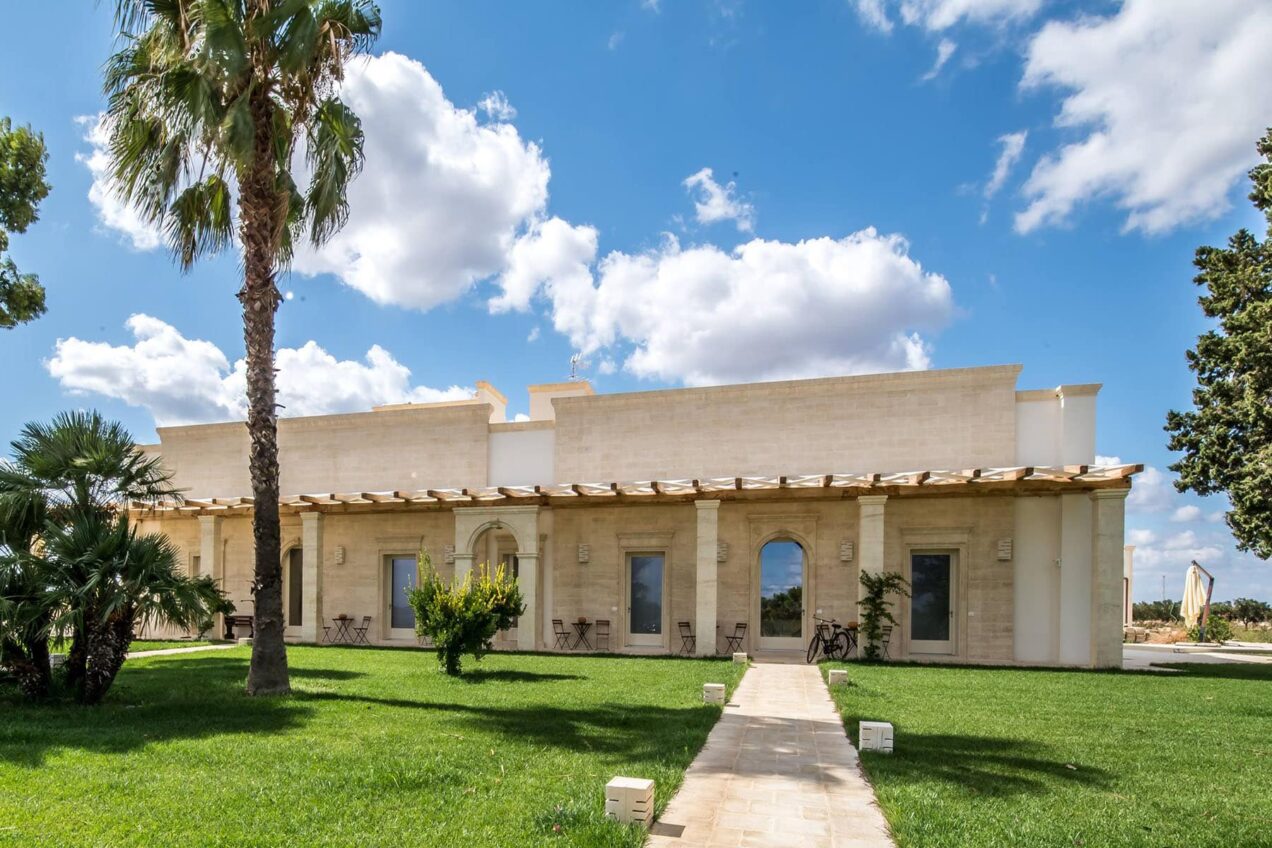 Modern architectural home with a landscaped front yard, featuring palm trees and a pathway leading to the entrance, under a partly cloudy blue sky. | Only on SopranoVillas