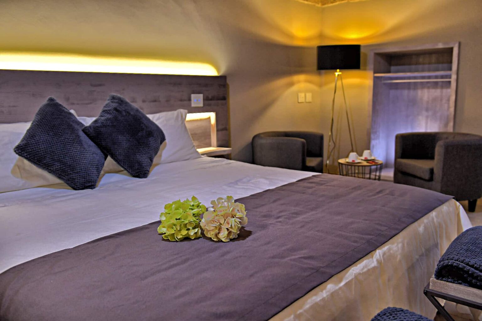 Modern hotel room with a king-sized bed adorned with decorative pillows, a cozy seating area, and elegant lighting, creating a relaxing atmosphere. Floral arrangements enhance the decor, making it inviting for guests. | Only on SopranoVillas
