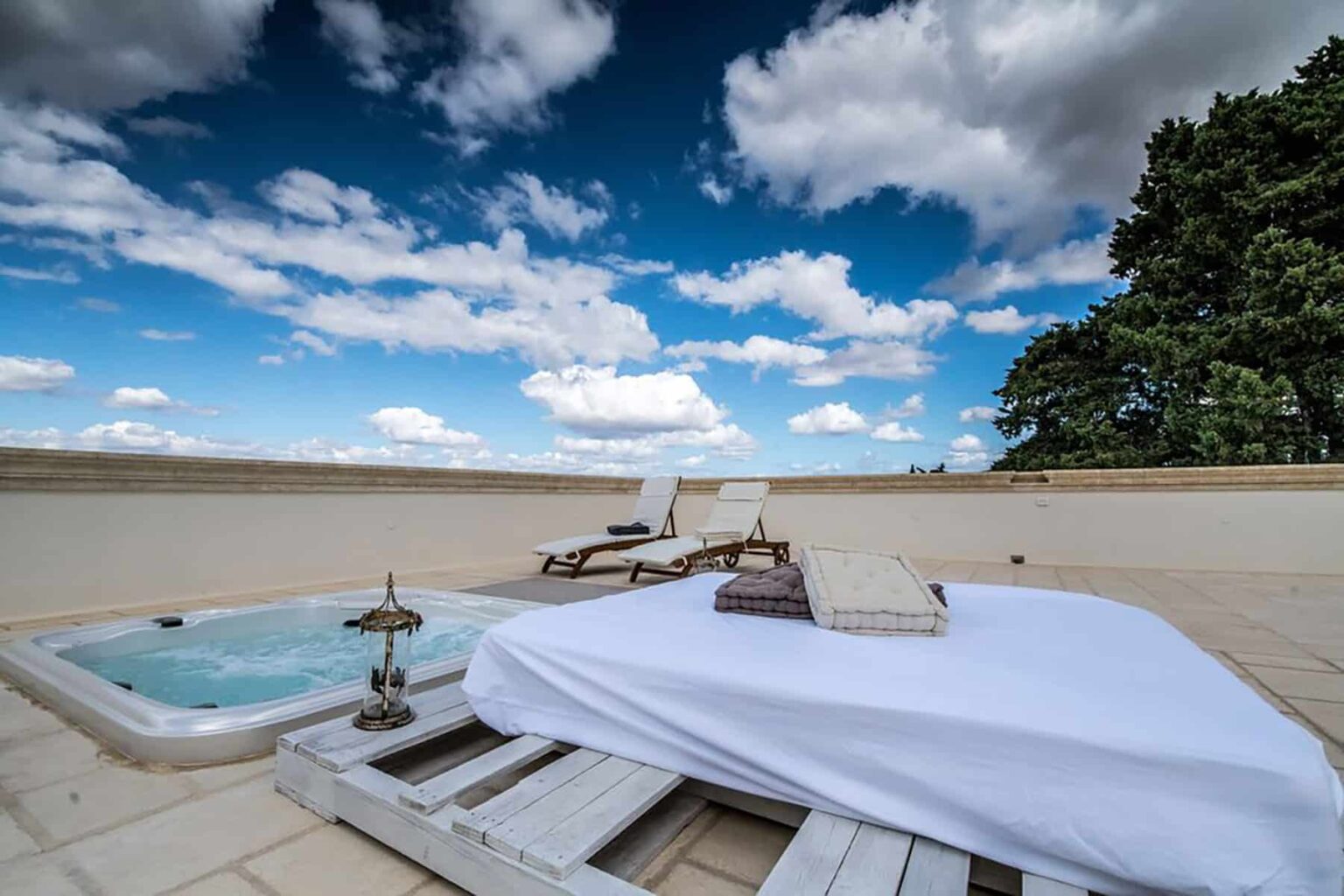Outdoor relaxation area featuring a hot tub and a sun lounger under a bright blue sky with fluffy clouds, surrounded by greenery. Ideal for leisure and wellness activities. | Only on SopranoVillas