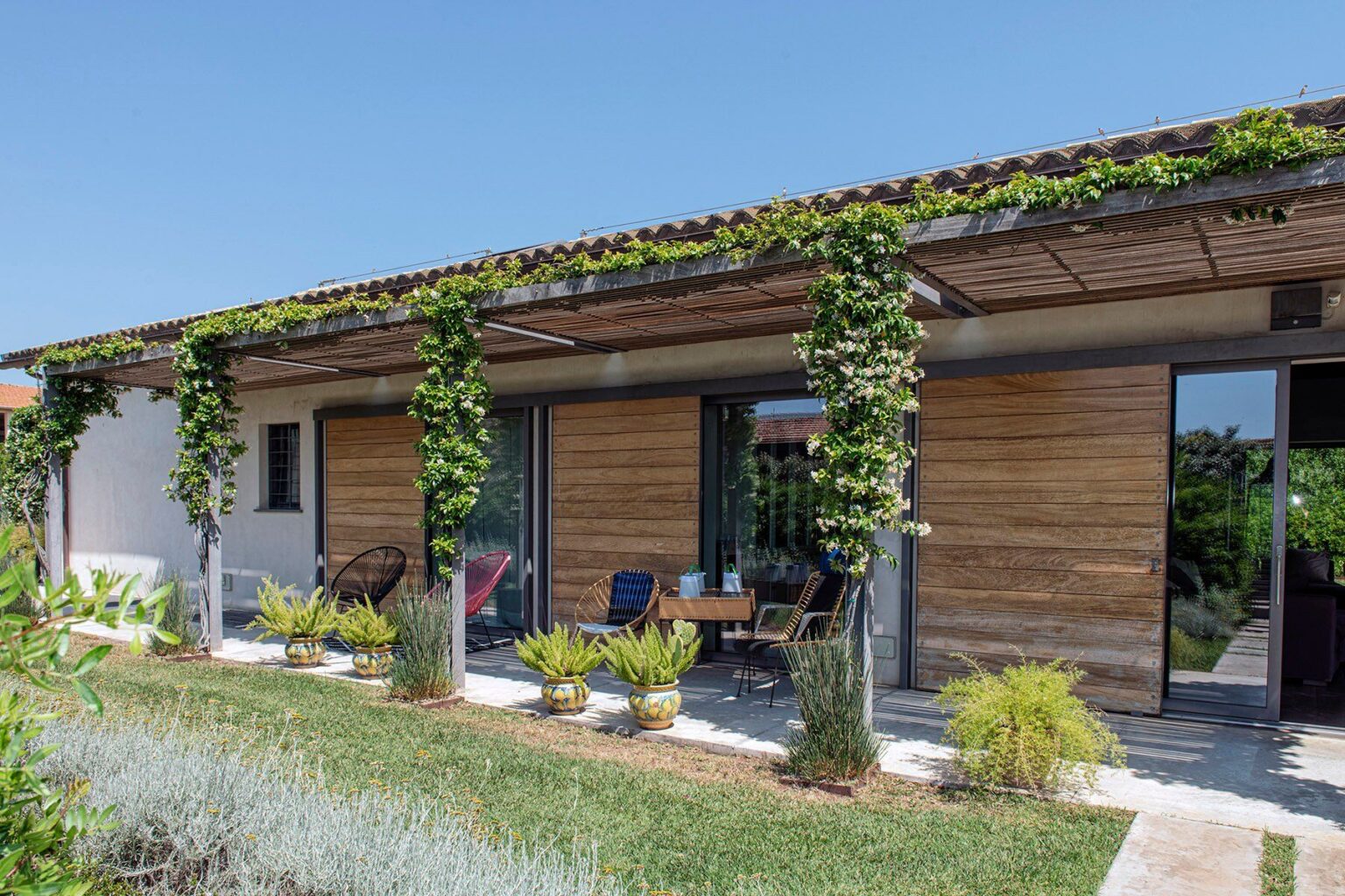 Modern eco-friendly house with wooden exterior and lush greenery, featuring large windows and a shaded patio area, surrounded by landscaped gardens. | Only on SopranoVillas