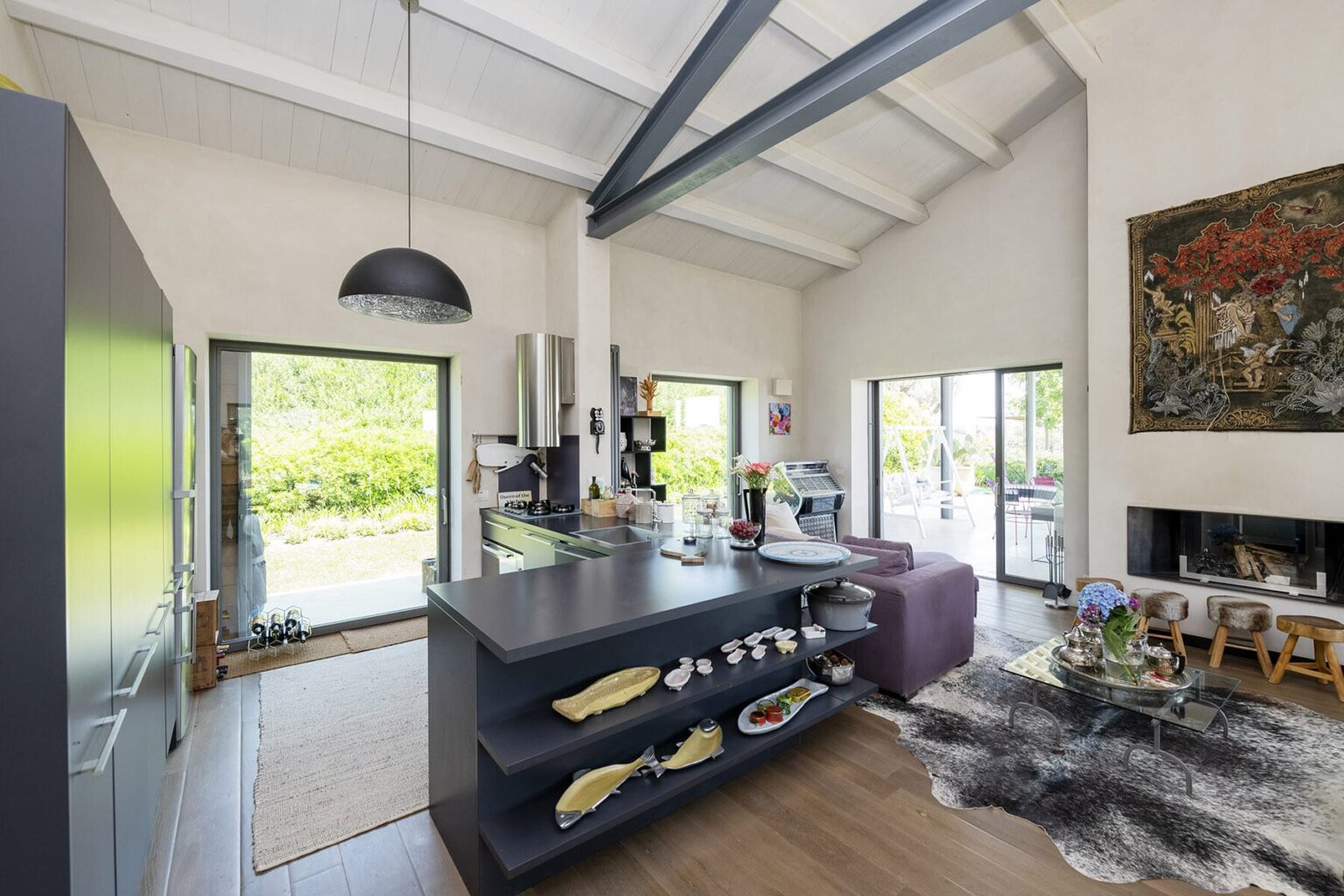 Modern open-concept kitchen and living room with high ceilings, featuring a sleek kitchen island, comfortable seating area, and large windows providing natural light and garden views. | Only on SopranoVillas