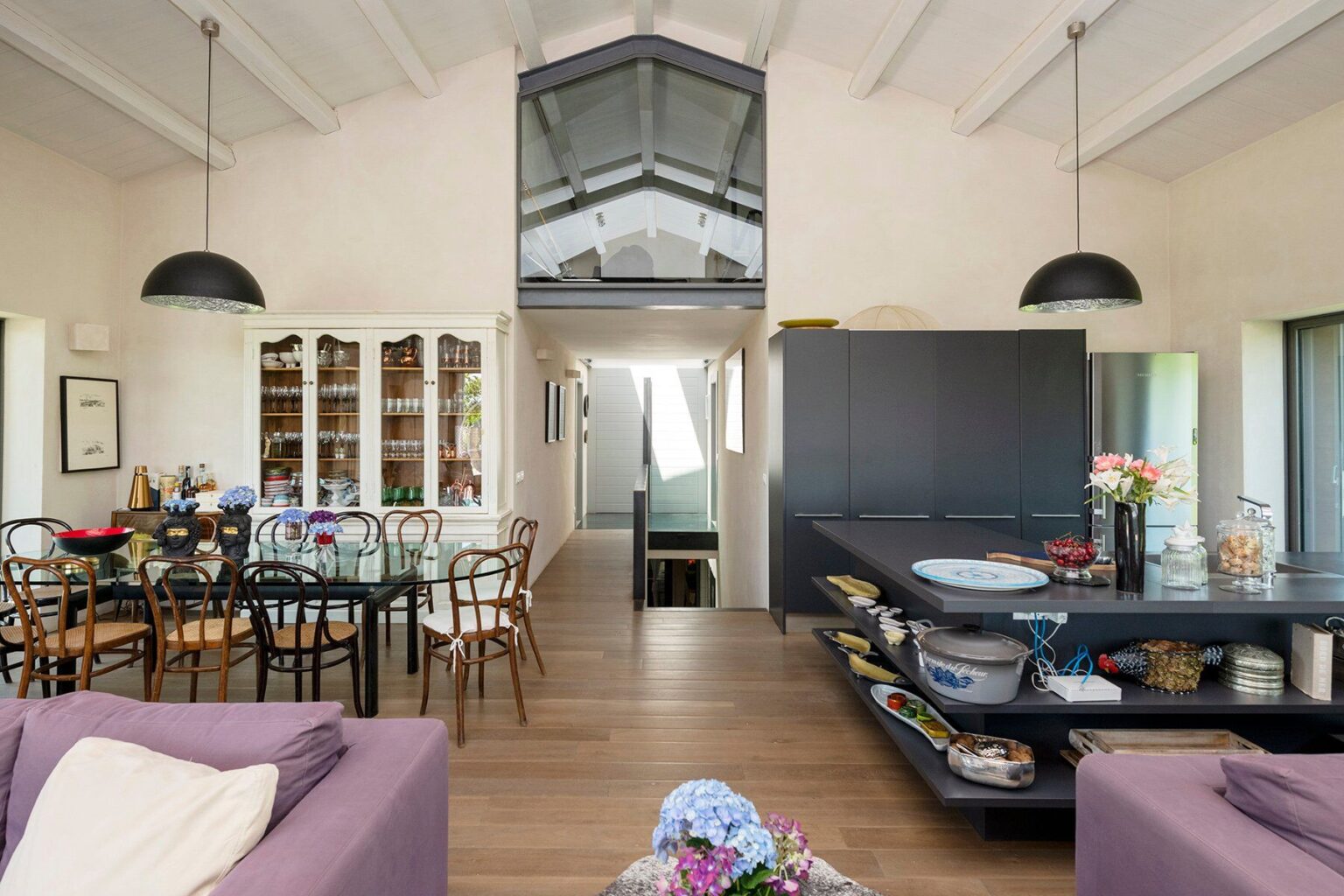 Modern open-plan living space featuring a stylish kitchen island, dining area with a glass table, and wooden flooring. The room is bright with high ceilings and large windows, showcasing a contemporary design aesthetic. | Only on SopranoVillas