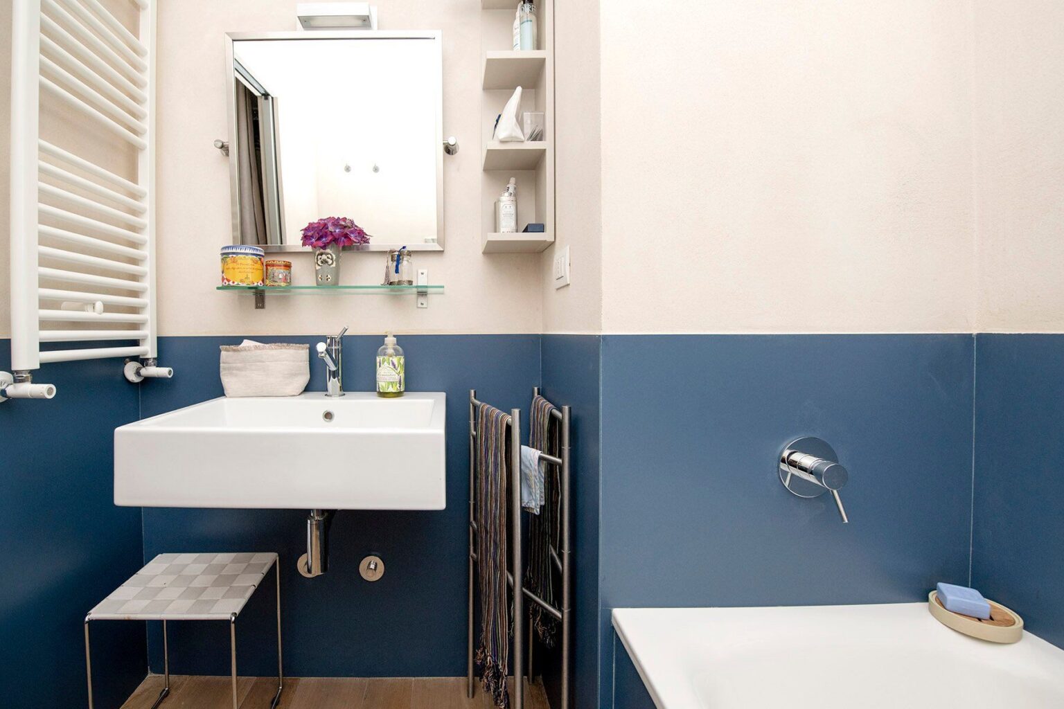 Modern bathroom interior featuring a sleek white sink, a wall-mounted mirror, and blue accent walls. The space includes a minimalist shelf for toiletries, a stylish towel rack, and a bathtub, creating a functional and aesthetically pleasing design. | Only on SopranoVillas