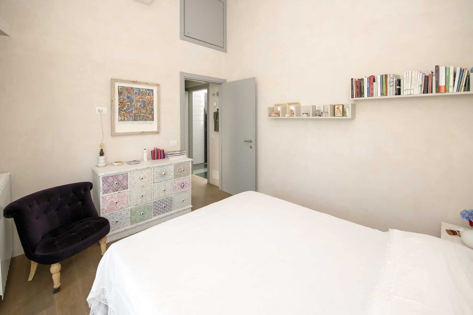 Cozy bedroom featuring a white bedspread, a stylish black armchair, and a wall shelf with books. The room has light-colored walls and a door leading to a bathroom, creating a welcoming and serene atmosphere. | Only on SopranoVillas