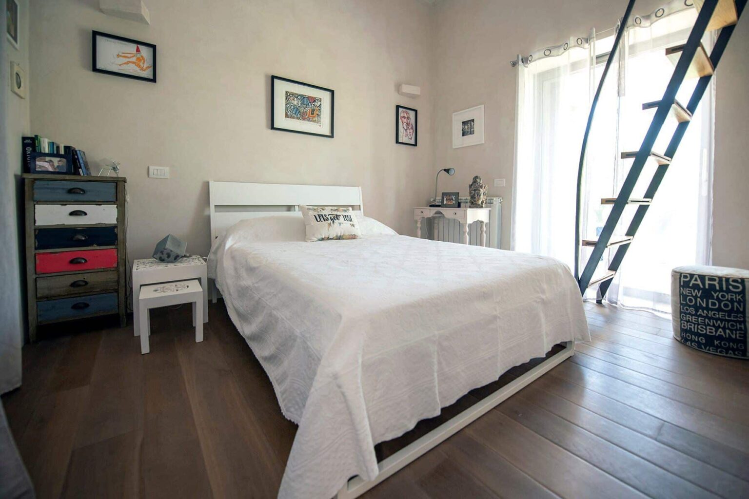 Cozy bedroom featuring a white bed with a textured blanket, a small bedside table, and a colorful dresser. Natural light streams through large windows, enhancing the inviting atmosphere. A decorative ladder leans against the wall, adding a unique touch to the room's design. | Only on SopranoVillas
