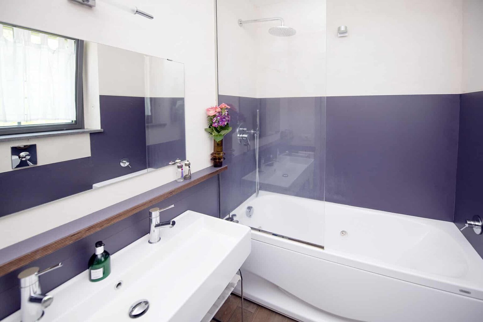 Modern bathroom featuring a sleek design with a bathtub, glass shower enclosure, and a stylish sink with a large mirror. The walls are painted in a calming purple hue, complemented by decorative flowers and toiletries on display. | Only on SopranoVillas