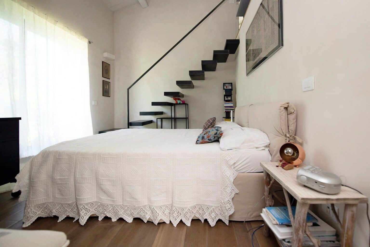 Cozy bedroom featuring a large bed with a decorative white bedspread, a bedside table with a lamp, and a staircase leading to an upper level. Natural light streams in through large windows, enhancing the inviting atmosphere. | Only on SopranoVillas