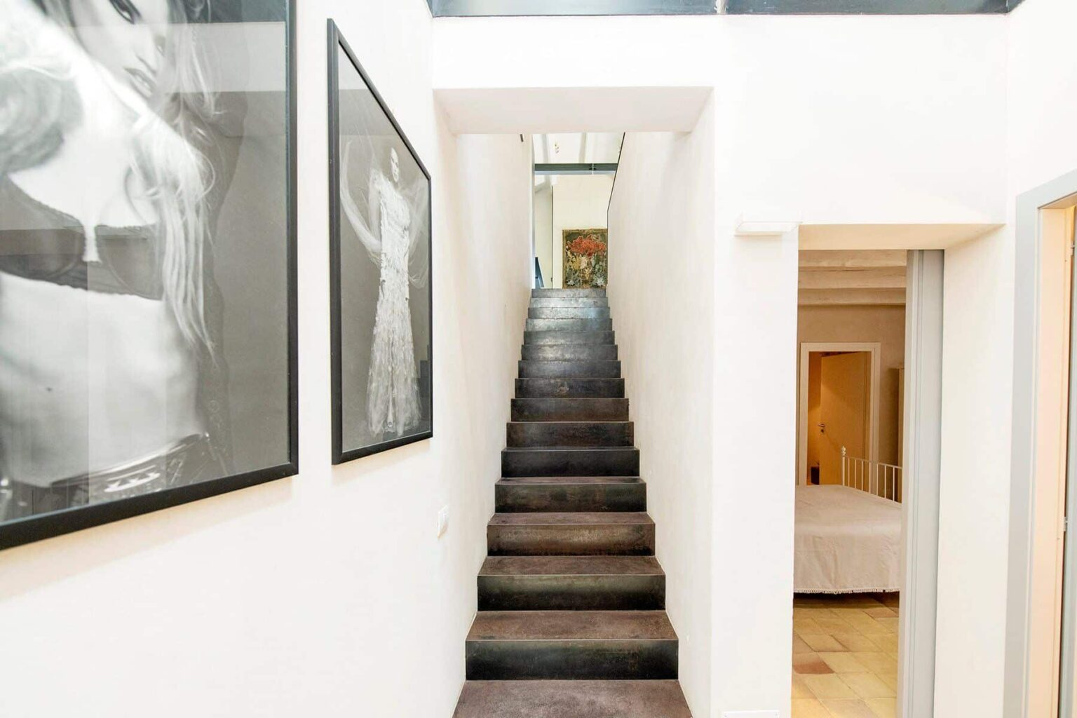 Modern interior featuring a staircase leading to an upper level, flanked by contemporary artwork on the walls. The hallway is well-lit, showcasing a minimalist design with white walls and wooden flooring. | Only on SopranoVillas