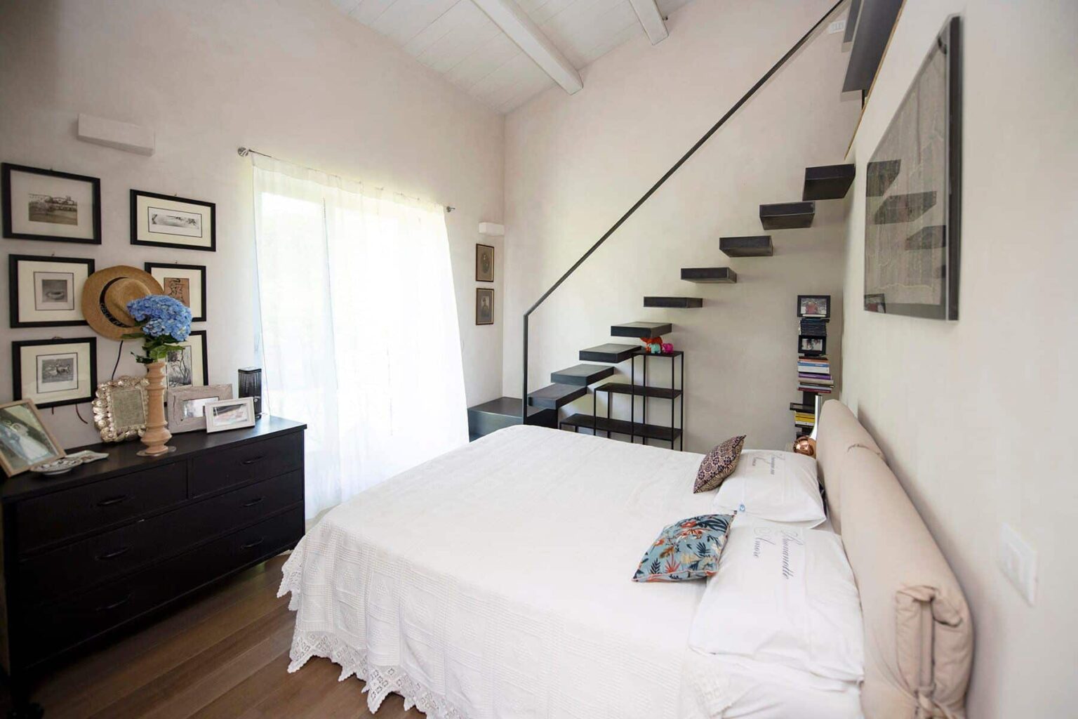 Cozy bedroom featuring a queen-sized bed with a light duvet, decorative pillows, and a stylish nightstand. The room includes a modern staircase leading to an upper level, large windows with sheer curtains allowing natural light, and framed artwork on the walls, creating a warm and inviting atmosphere. | Only on SopranoVillas