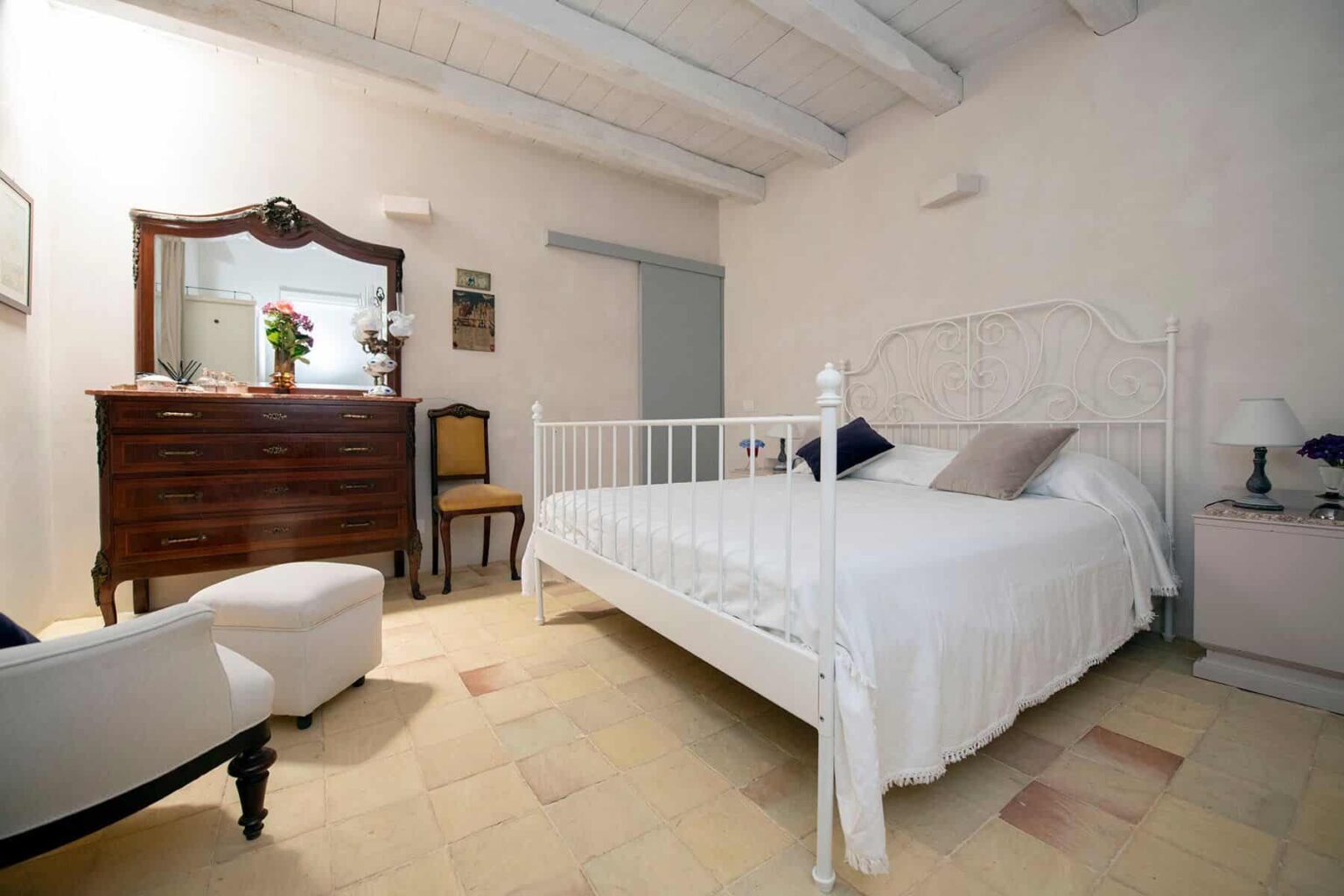 Cozy bedroom featuring a white metal bed, wooden dresser, and a comfortable armchair, with soft lighting and rustic decor. | Only on SopranoVillas