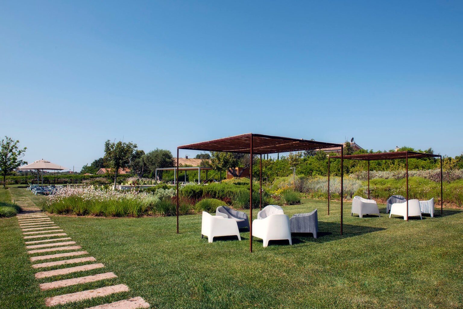 Outdoor seating area with white chairs under a shaded pergola, surrounded by lush greenery and a clear blue sky, ideal for relaxation and gatherings. | Only on SopranoVillas