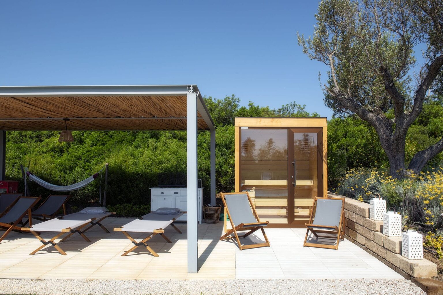 Modern outdoor relaxation area featuring a covered patio with lounge chairs, a hammock, and a wooden sauna surrounded by greenery. Ideal for leisure and wellness activities in a serene setting. | Only on SopranoVillas