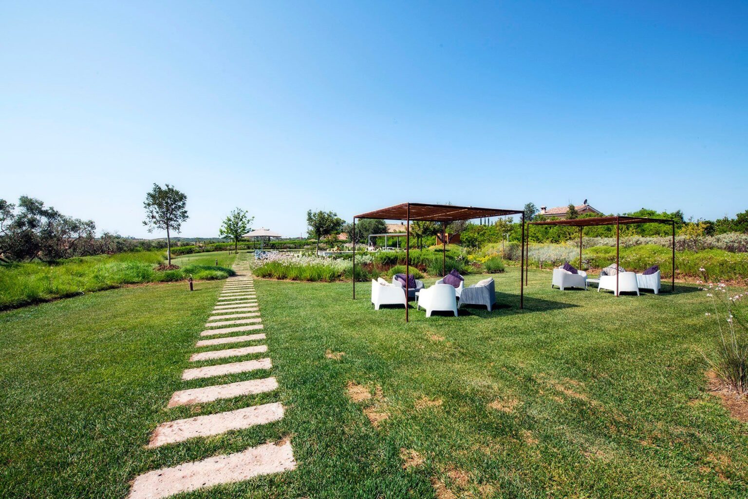 Lush green garden with a stone pathway leading to shaded seating areas, surrounded by vibrant flowers and clear blue sky. Ideal for relaxation and outdoor gatherings. | Only on SopranoVillas