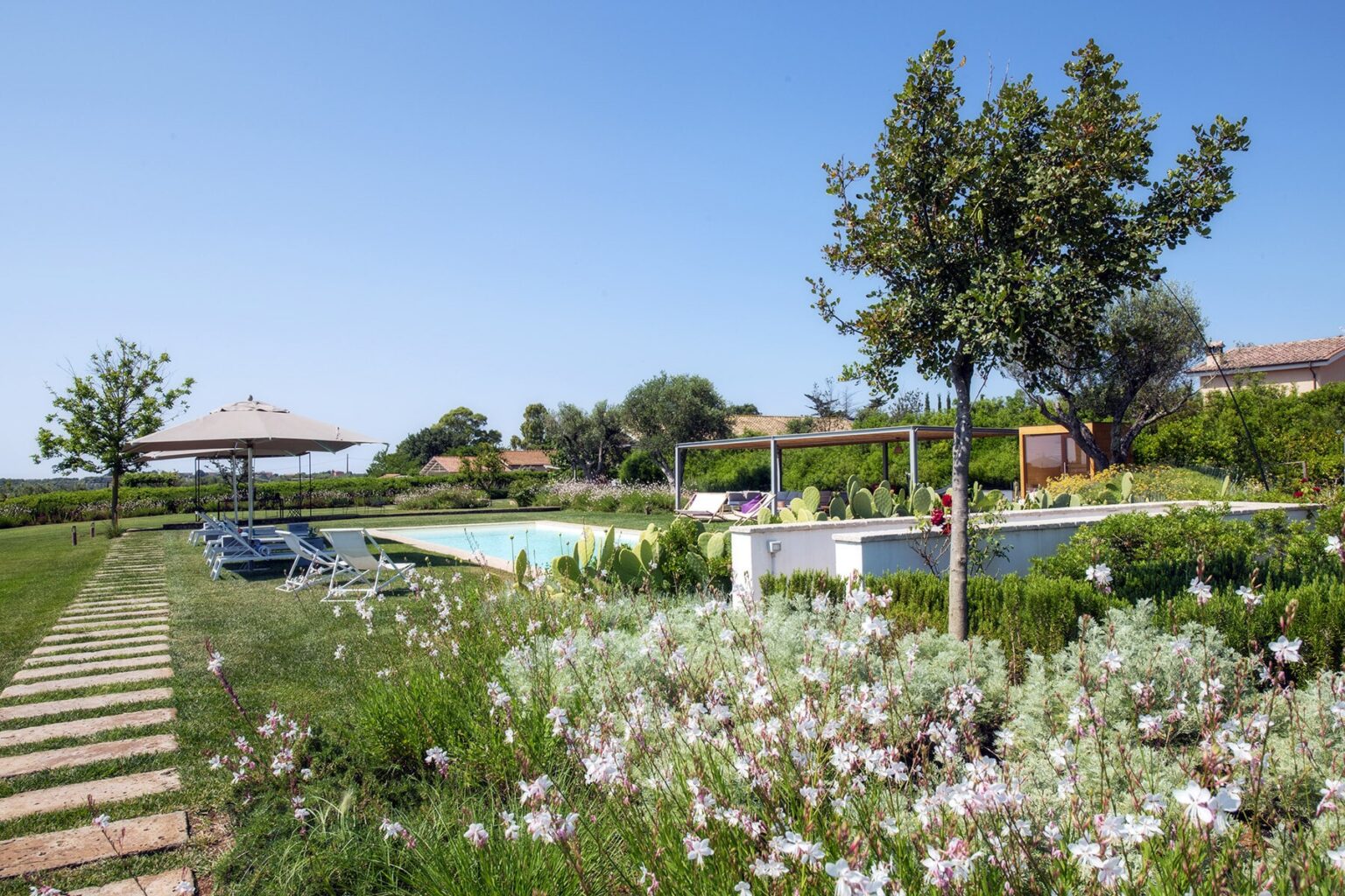 Lush garden with a swimming pool and lounge chairs, surrounded by flowering plants and trees under a clear blue sky. | Only on SopranoVillas