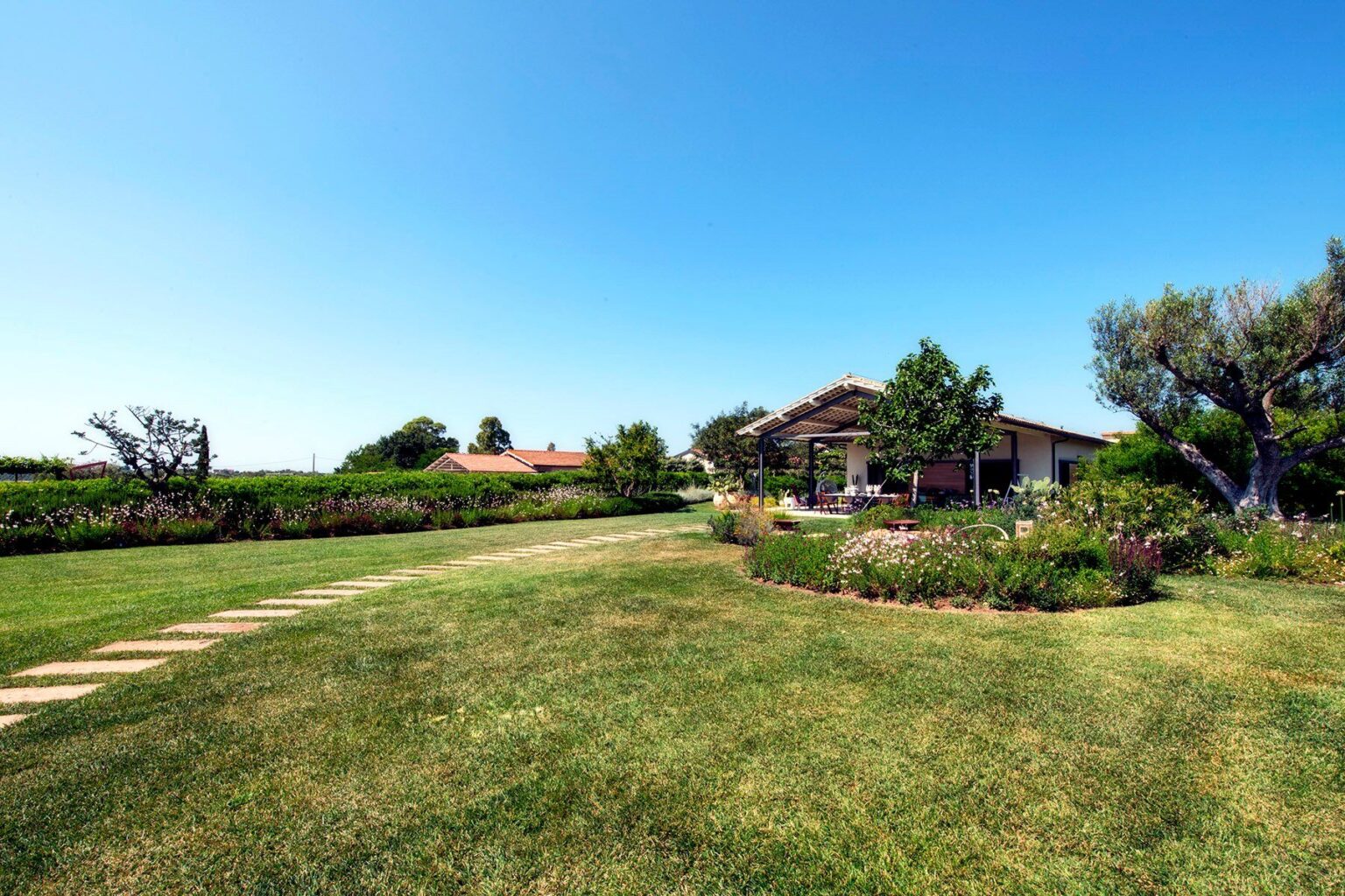 Scenic landscape featuring a well-maintained garden and a charming house surrounded by lush greenery and clear blue skies, ideal for outdoor living and relaxation. | Only on SopranoVillas