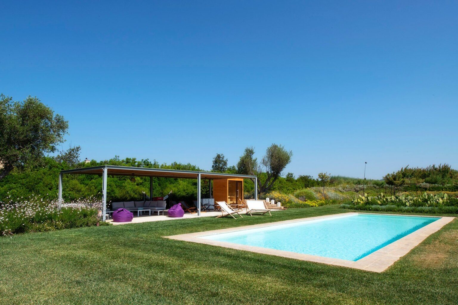 Modern outdoor pool surrounded by lush greenery and a shaded seating area with lounge chairs and bean bags under a clear blue sky. Ideal for relaxation and summer gatherings. | Only on SopranoVillas